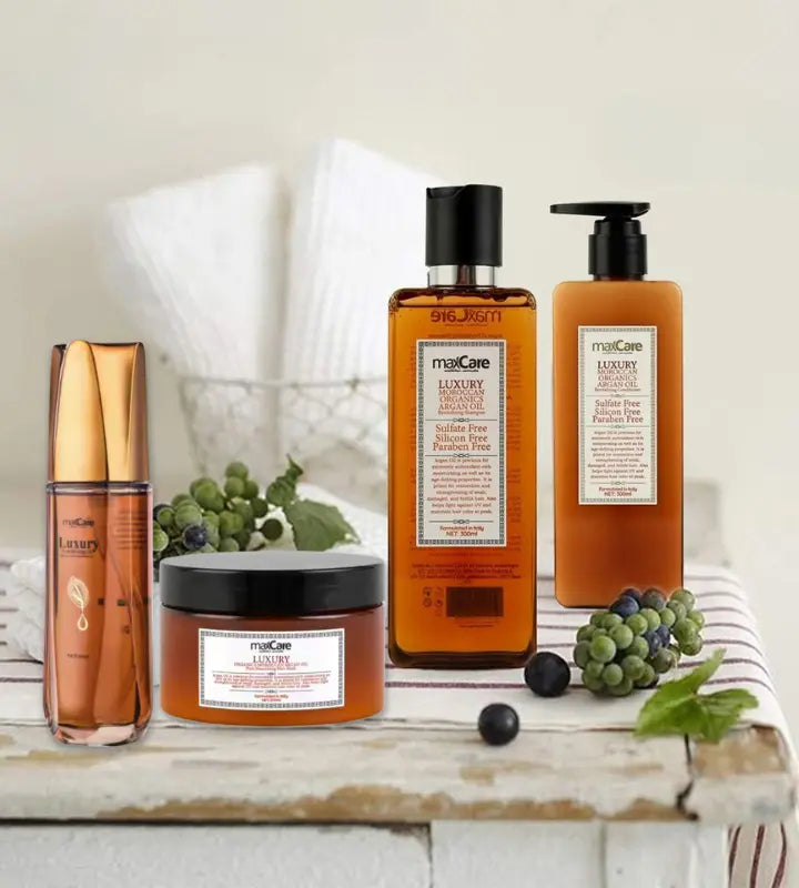 MaxCare Luxury Moroccan Argan Oil hair care collection displayed on a spa-style white wooden table with fresh berries and mint leaves. The set includes sulfate-free, silicone-free, and paraben-free shampoo, conditioner, hair mask, and oil serum, all in amber-toned packaging with a premium, natural aesthetic.
